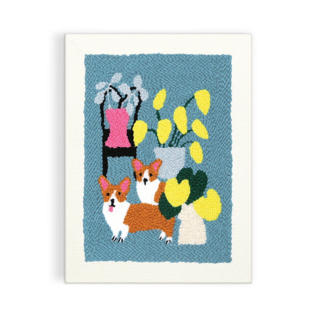 The Anne Bentley Corgi Friends Punch Needle Kit features an embroidered artwork of two brown and white corgis, a black chair with a pink cushion, and vases with colorful leaves on a blue background.