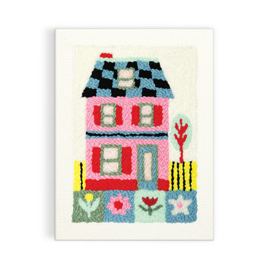 The Cozy Cottage Punch Needle Kit by Molly Egan features a vibrant embroidered pink house with a blue and black checkered roof, green door, windows, flowers, and a tree on a white background.