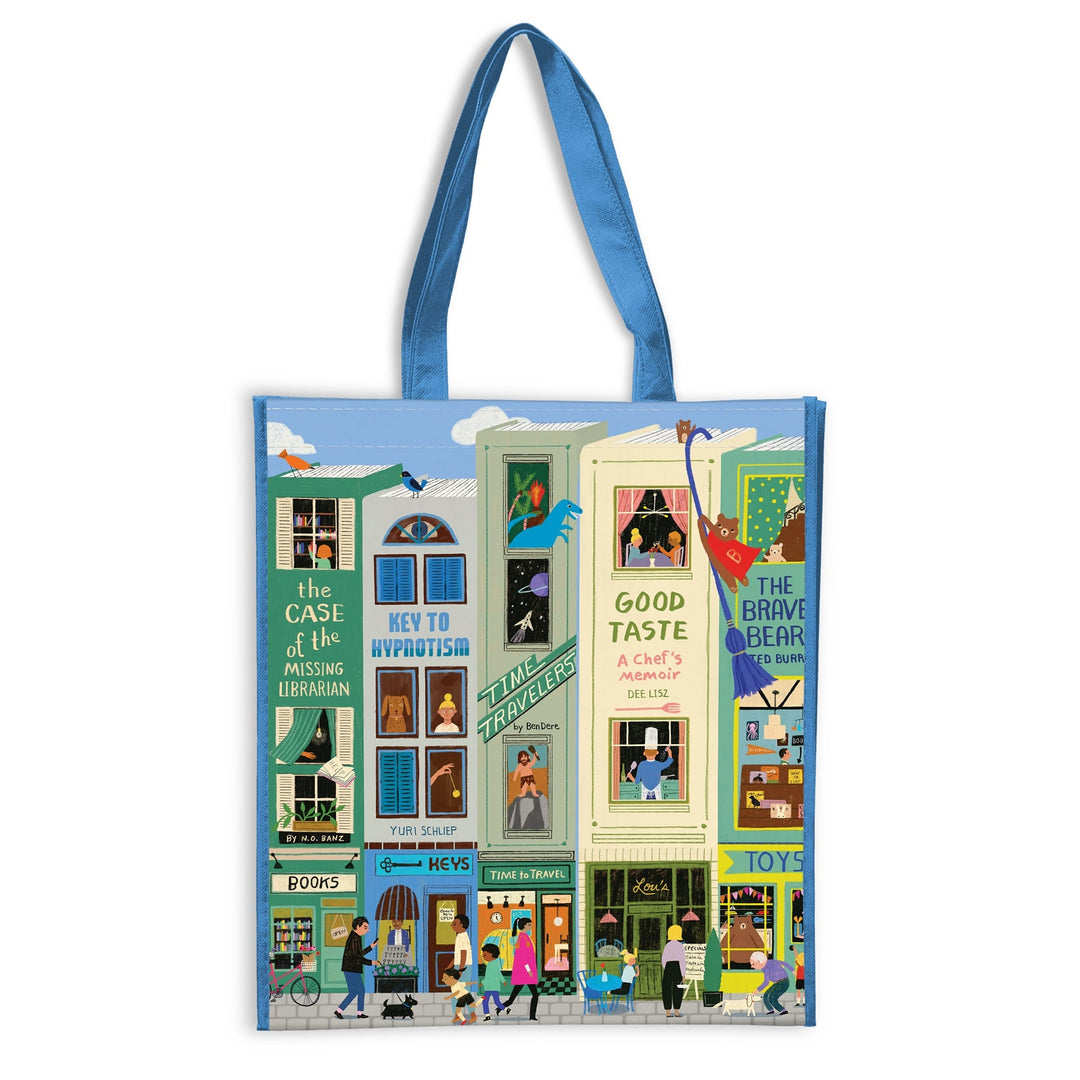 The Main Street Books Reusable Shopping Bag by Anne Bentley features blue handles and vibrant artwork of storefronts, apartments, a bookshop, toy store, café, plus people and animals bringing the lively street scene to life.