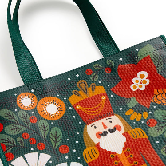 The Merry Nutcracker Reusable Shopping Bag by Sandra Hutter features festive holiday artwork with a nutcracker, red flowers, pinecones, and green foliage on a dark green background with scattered white dots and green handles.