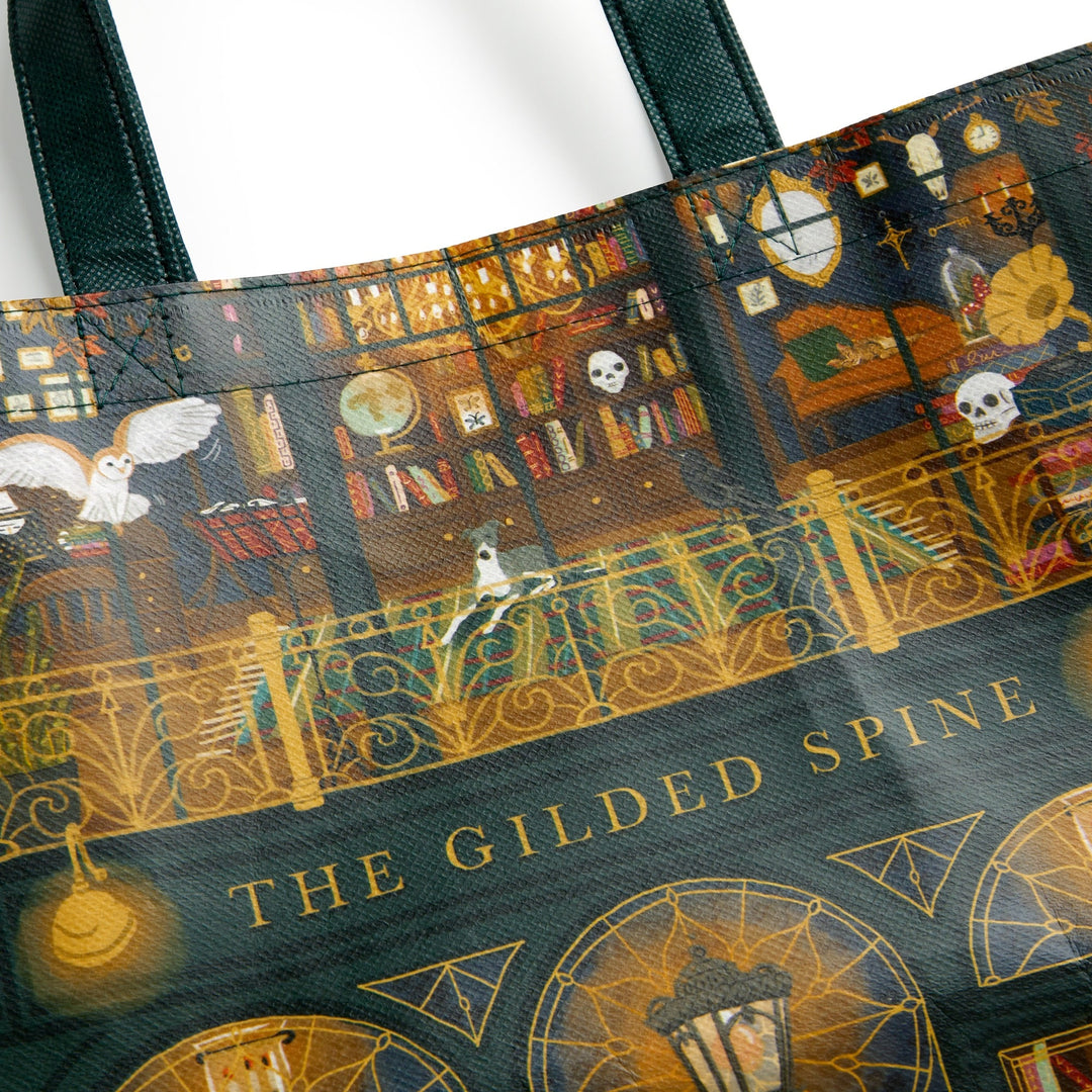 A close-up of the Sally Wilson Gilded Spine Reusable Shopping Bag, showcasing a whimsical library scene with bookshelves, magical objects, an owl, golden details, and "THE GILDED SPINE" in gold at the bottom.