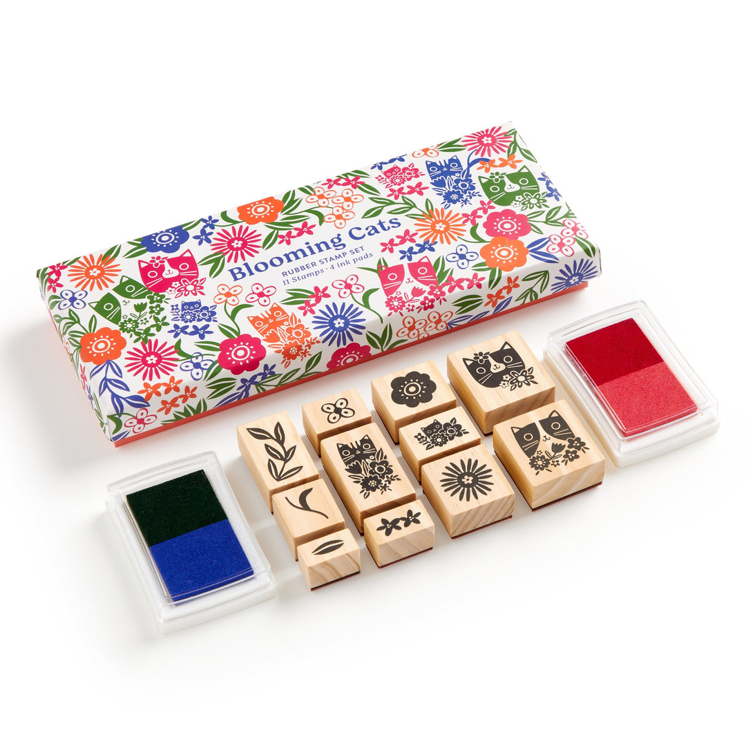 A Tara Lilly Blooming Cats Rubber Stamp Set, featuring nine wooden stamps with botanical and cat designs plus red and blue-green ink pads, comes in a colorful box. The ink pads sit on either side of the vibrant packaging.