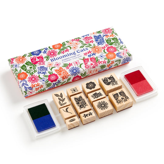 A Tara Lilly Blooming Cats Rubber Stamp Set, featuring nine wooden stamps with botanical and cat designs plus red and blue-green ink pads, comes in a colorful box. The ink pads sit on either side of the vibrant packaging.