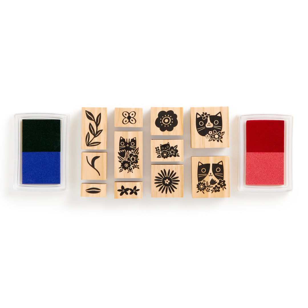 The Tara Lilly Blooming Cats Rubber Stamp Set includes eleven wooden stamps with cat and floral designs, arranged in a row. A blue and black ink pad is on the left, and a red ink pad is on the right.