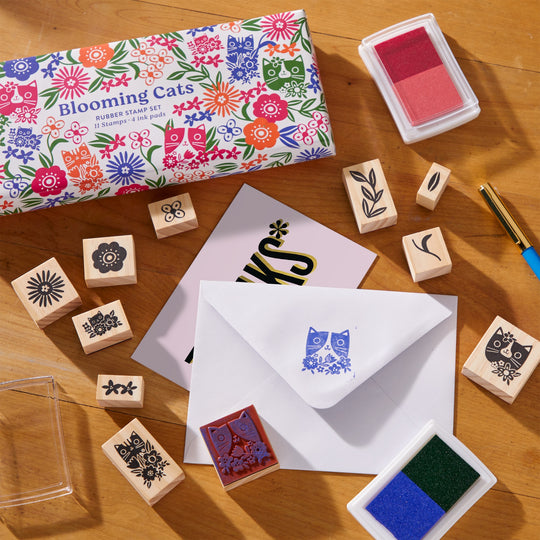 A colorful box labeled Blooming Cats Rubber Stamp Set by Tara Lilly sits on a wooden table, showcasing feline and botanical designs on rubber stamps, ink pads, a pen, plus a card and stamped envelope.