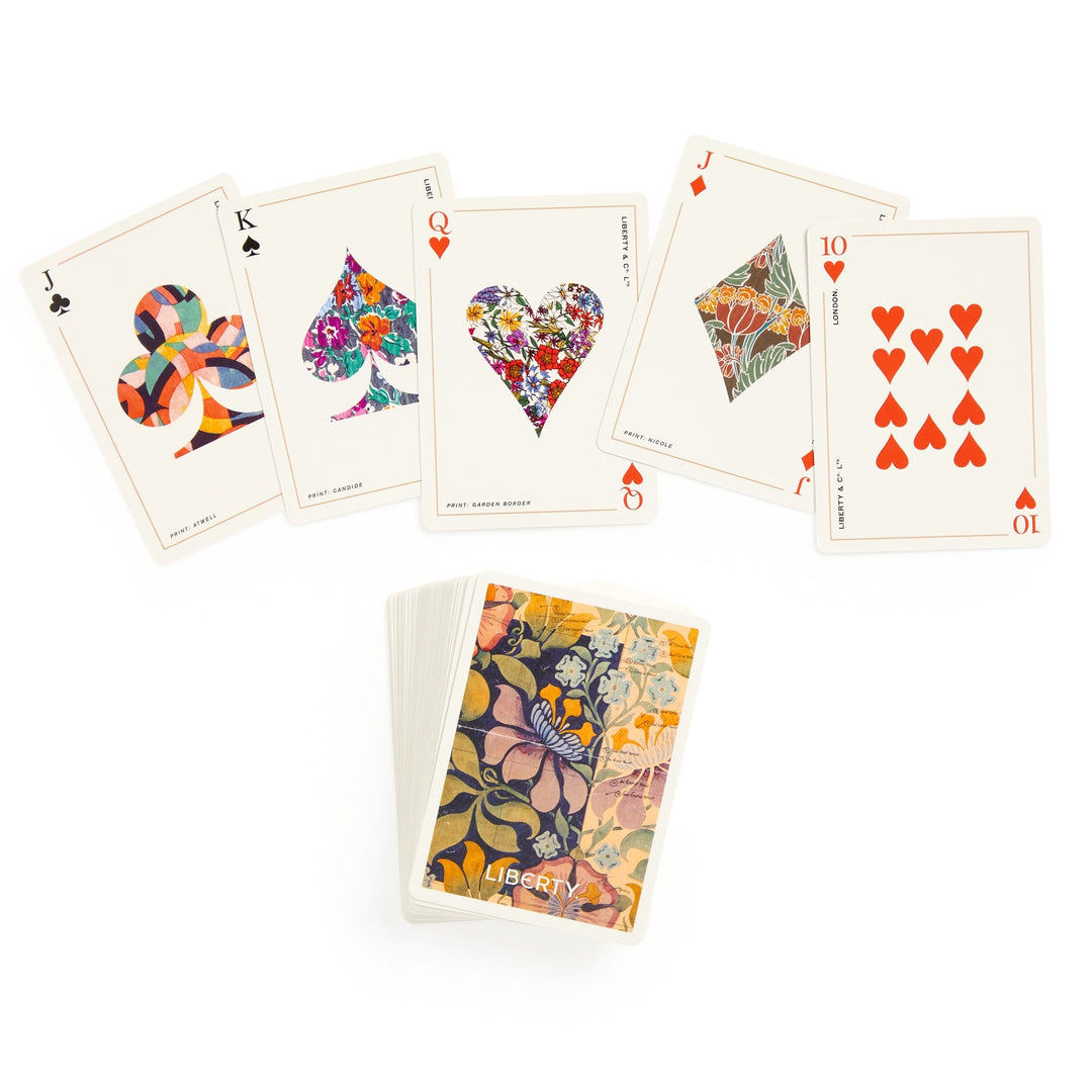 A spread of Liberty London playing cards from the Liberty Archive Playing Card Deck displays unique, colorful patterns on the Jack of Clubs, King and Queen of Spades, Queen and Ten of Hearts. The deck lies stacked face down beneath them.