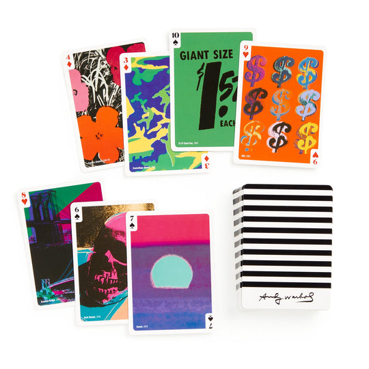 The Warhol Pop Art Playing Card Deck by the Andy Warhol Foundation For The Visual Arts features premium cards with vibrant Pop Art images—flowers, dollar signs, a skull, and a sunset—packaged in a striped box labeled Andy Warhol.
