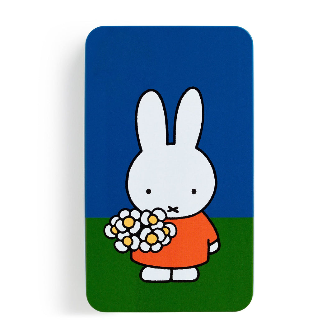 The Miffy Sticker Tin by Miffy features a cartoon white bunny with long ears, wearing an orange dress and holding white daisies on green grass beneath a simple, colorful blue sky.