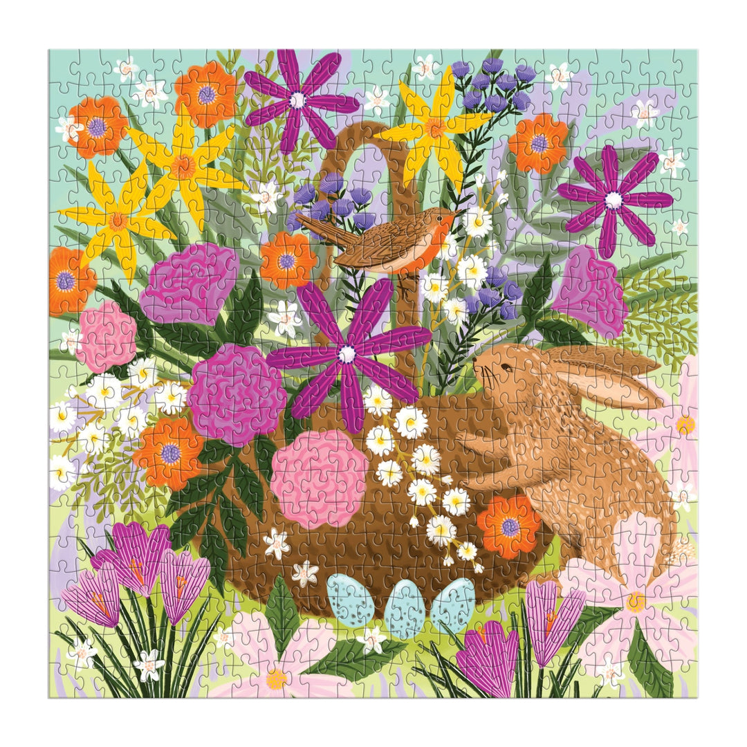 The Good Puzzle Co. Basket Blooms 500pc Puzzle by Lee Foster-Wilson features a spring scene: colorful flowers, a bird on a nest, blue eggs in grass near a brown bunny, all set against a light blue and pastel background.