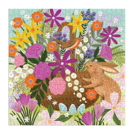 The Good Puzzle Co. Basket Blooms 500pc Puzzle by Lee Foster-Wilson features a spring scene: colorful flowers, a bird on a nest, blue eggs in grass near a brown bunny, all set against a light blue and pastel background.
