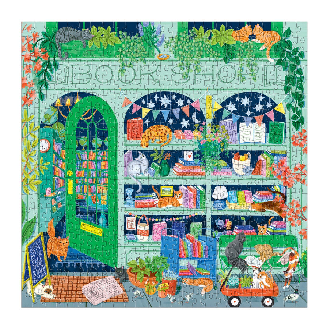 The Good Puzzle Co. Bookshop Cats 500pc Puzzle by Lee Foster-Wilson features a cozy bookshop exterior with cats, books, plants, and a chalkboard sign. Eco-friendly pieces reveal playful felines lounging among shelves and a wagon of books outside.