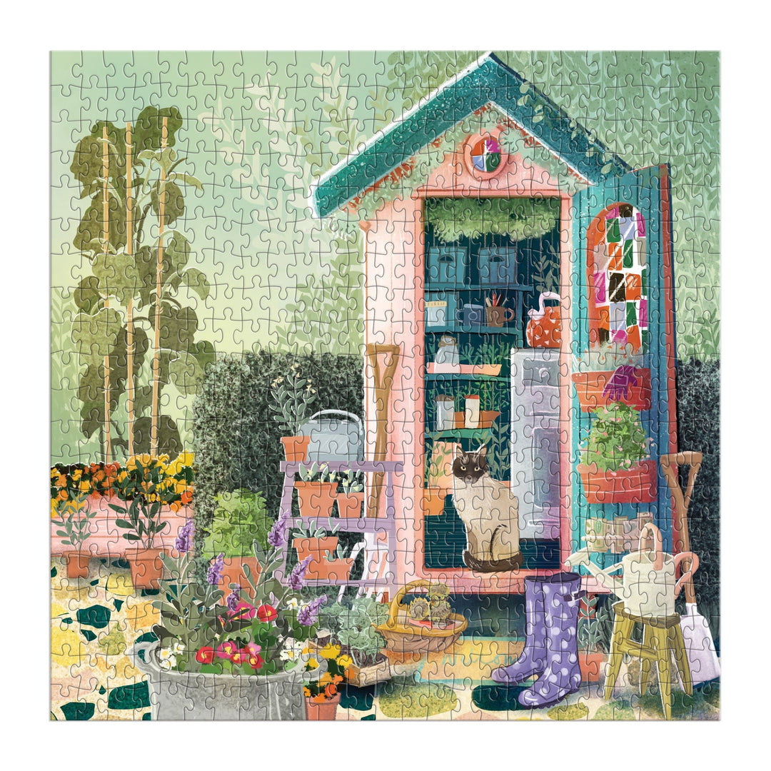The Good Puzzle Co. Gardeners Nook 500pc Puzzle by Bex Parkin features a colorful garden shed with a cat inside, surrounded by blooming flowers, gardening tools, rubber boots, and potted plants in lush greenery.