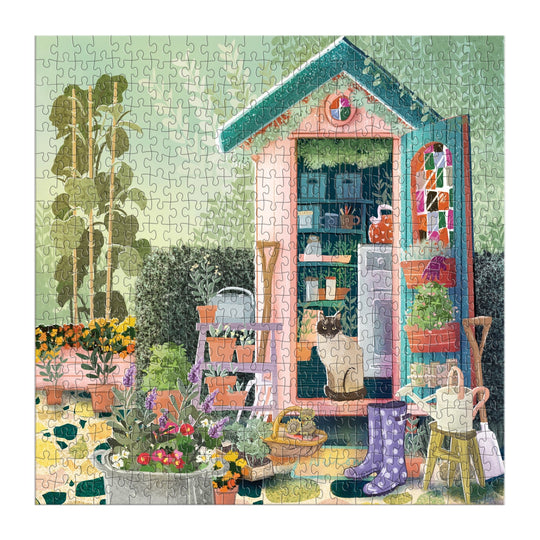 The Good Puzzle Co. Gardeners Nook 500pc Puzzle by Bex Parkin features a colorful garden shed with a cat inside, surrounded by blooming flowers, gardening tools, rubber boots, and potted plants in lush greenery.