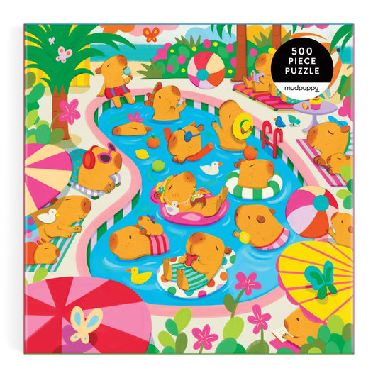 The Valerie Nelson Capybara Pool Day 500 Piece Family Puzzle features a lively scene of capybaras relaxing in a pool, floating on rings, and playing with rubber ducks, surrounded by umbrellas and plants—perfect for family fun.