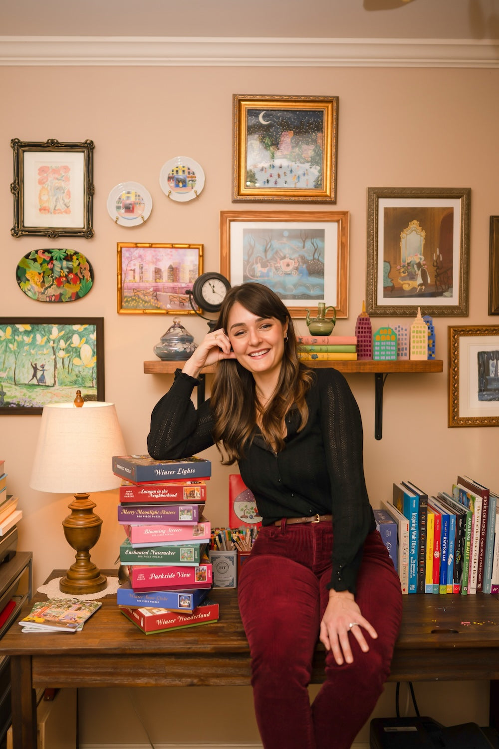 A smiling woman with long brown hair sits on a desk beside a stack of books, surrounded by framed art and plates on a beige wall. Books and a lamp are on the desk and shelves. The setting is cozy and colorful.